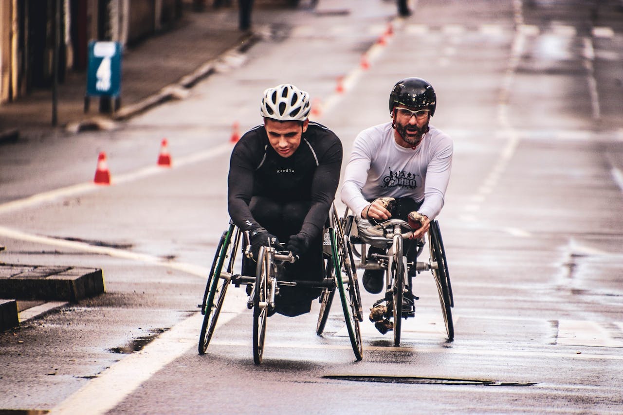 our-story-01 Two men compete in a wheelchair race on a wet urban street, showcasing endurance and teamwork.