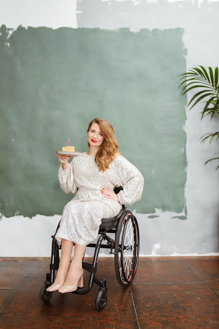 Elegant woman with red lipstick in a wheelchair holding a birthday cake indoors.