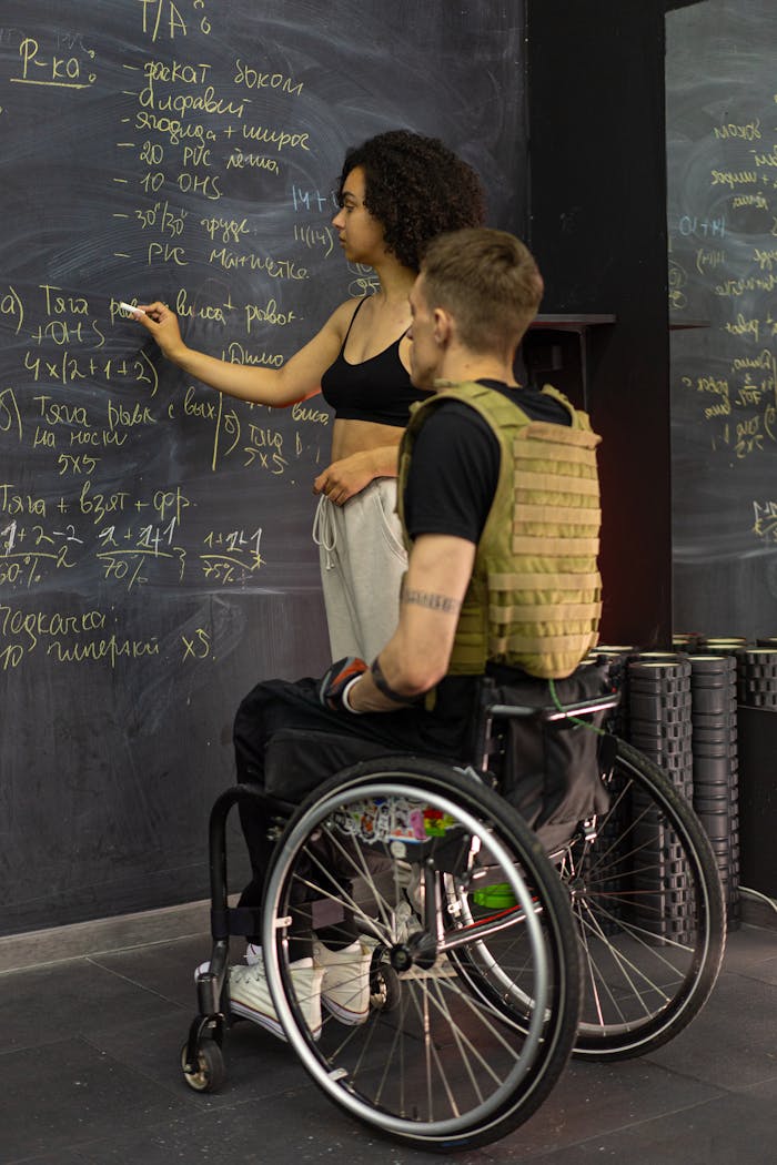 Teacher instructing student in a wheelchair at blackboard. Collaborative learning environment indoors.